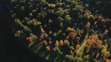 Bird's-eye-view-of-a-narrow-gravel-pathway-and-a-asphalt-road-through-a-colorful-dense-forest-in-the-rural-area-of-Litchfield-county,-Connecticut,-USA-on-a-sunny-morning