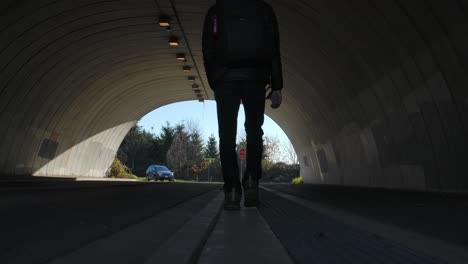 Silueta-Retroiluminada-De-Una-Persona-Caminando-Por-Un-Túnel,-Un-Coche-Acercándose-Al-Fondo,-Luz-Del-Día