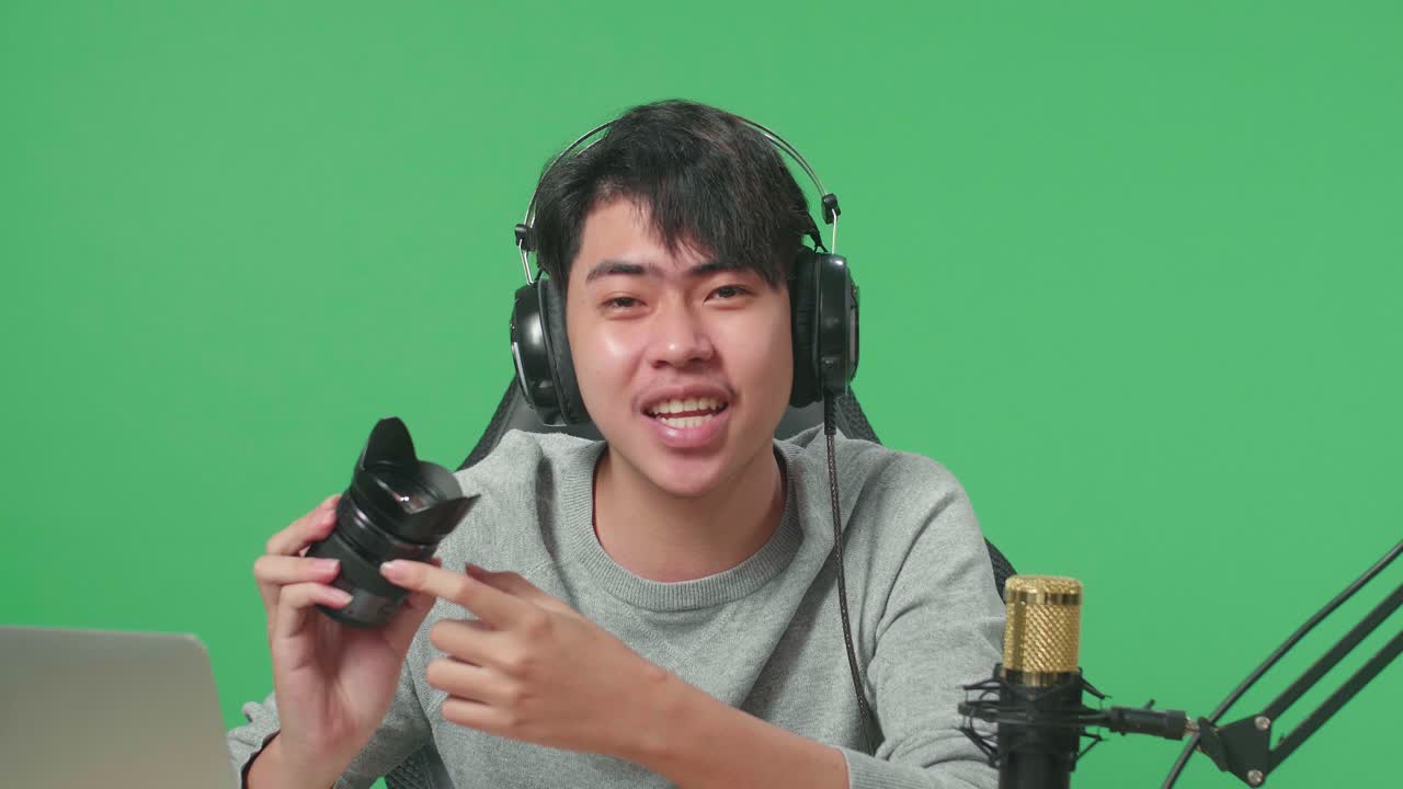 Close Up Of Smiling Asian Man With Computer Reviewing Camera Len While Sitting In Front Of Green Screen Background