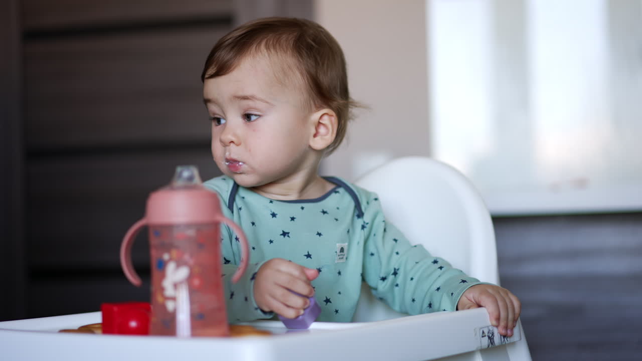 Cute adorable baby boy sitting in the feeding chair. Caucasian child with smeared face playing with a toy. Blurred backdrop.