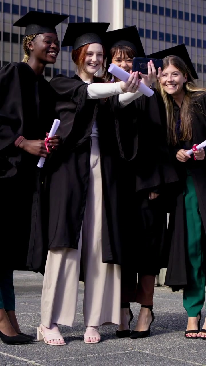 Vertical video: Fixing cap, red gowned grad pulling phone on plaza, taking selfie holding diploma