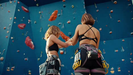 Rear-view-of-a-blonde-girl-installing-the-necessary-equipment-and-attaching-the-belay-to-herself-and-her-friend-before-her-fire-on-a-blue-climbing-wall-on-a-sunny-day-in-summer