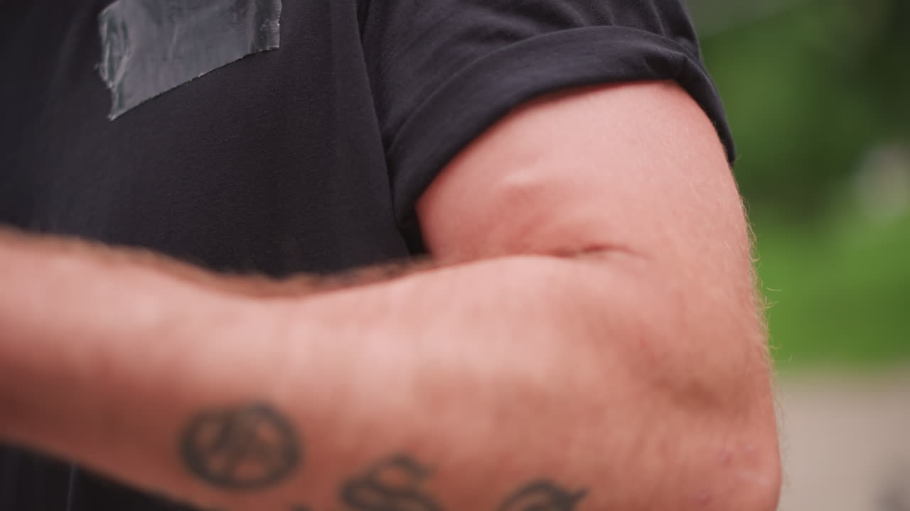 White Male Arm Detail With Rolled Sleeve Showcasing Bicep And Tattoo, Gym Enthusiast Flexing With Subtle Movement, Outdoor Park Background And Natural Light Highlighting Muscle Texture And Confident