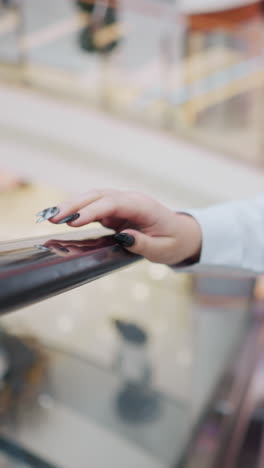woman with black polished nails gently tapping escalator handrail while descending, she wears a white blouse and jeans, soft-focus background captures a modern shopping mall with blurred shoppers