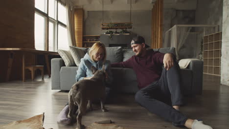 Couple-Sitting-On-The-Floor-Leaning-On-The-Sofa-Caressing-Their-Bulldog-Dog