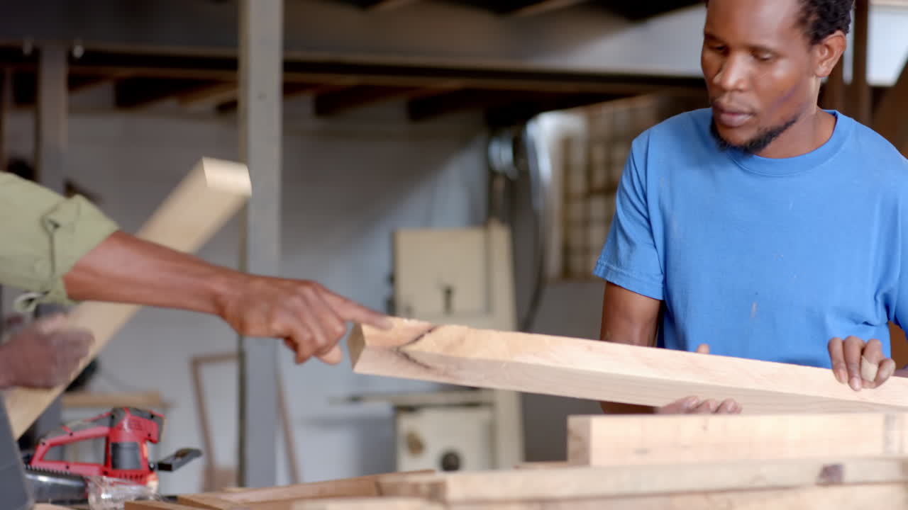 Inspecting wooden planks African American coworkers working in carpentry workshop, with power drill