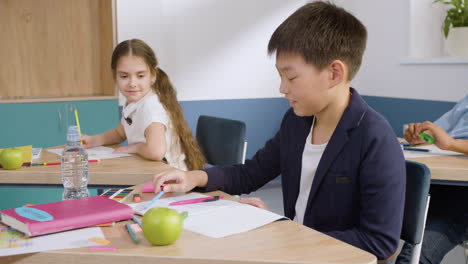 male student sitting at desk picks up a pencil from her classmate's desk