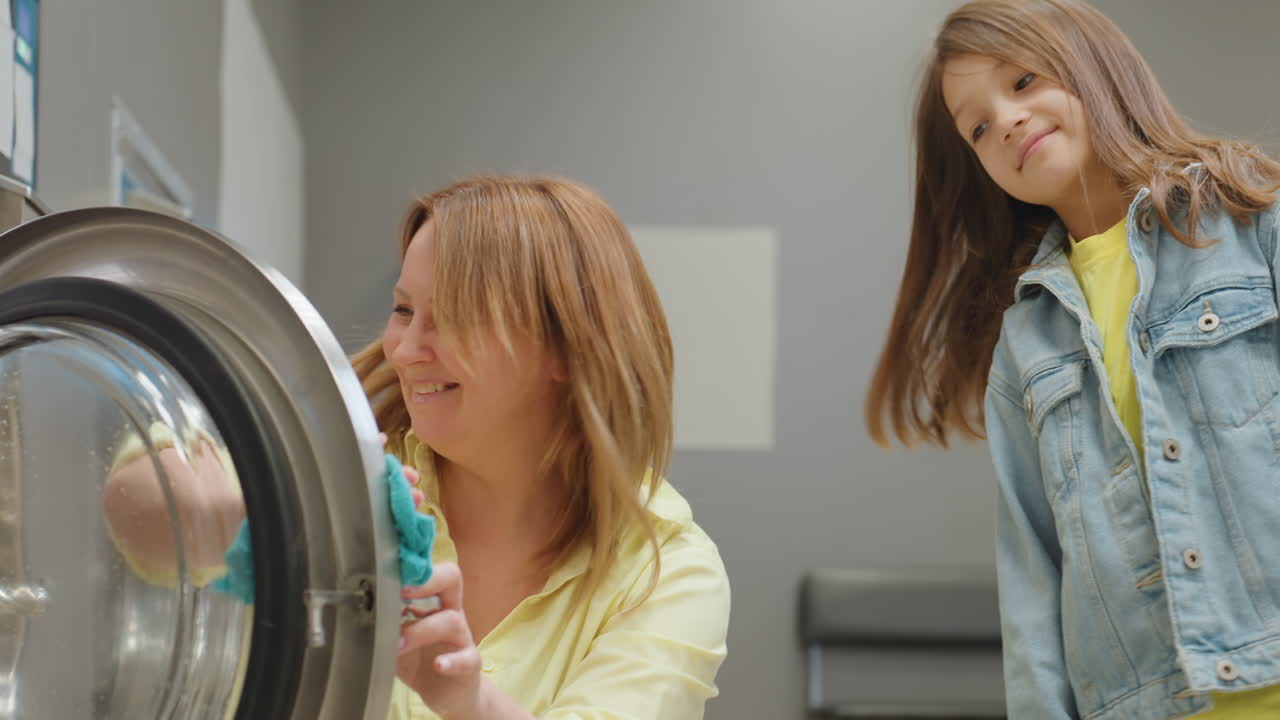 Joyful mother smiles warmly as toddler stands beside her while they enjoy playful bonding moment cleaning washing machine door with wet towel, creating cheerful teamwork atmosphere