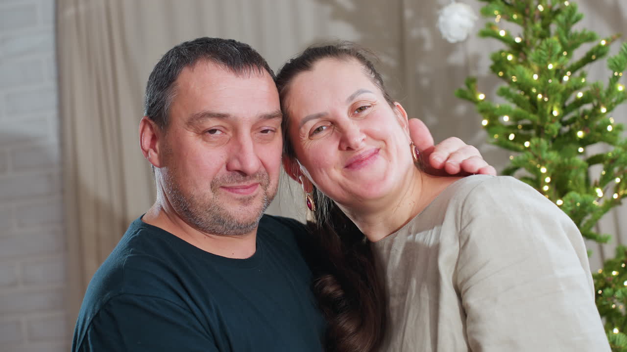 Husband and wife lovingly join foreheads and smile warmly as husband kisses wife's cheek in cozy room decorated with Christmas tree and twinkling lights, expressing deep affection, connection