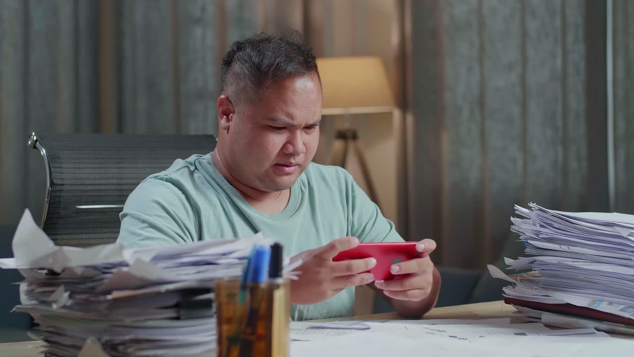 Primo piano di un uomo asiatico corpulento che è contrariato per aver perso una partita sullo smartphone dopo aver lavorato a dei documenti in ufficio.