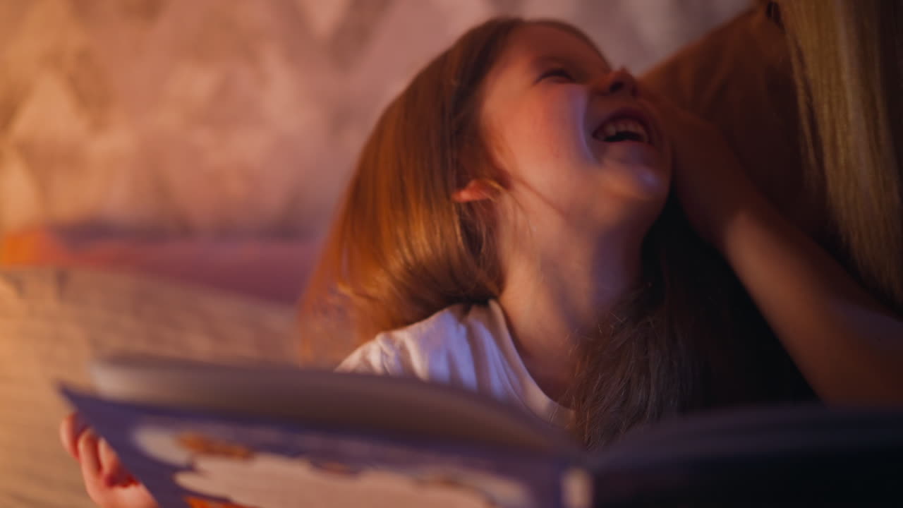 madre acaricia a la niña riendo con el libro de la cola de hadas en la cama