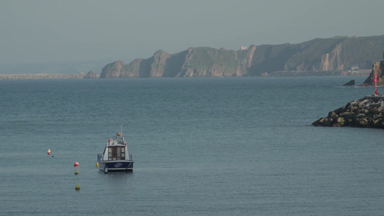 Peaceful coastal view of a small fishing boat floating on the Atlantic Ocean near Candás, Asturias, Spain. Distant cliffs and calm blue water create a scenic maritime landscape.