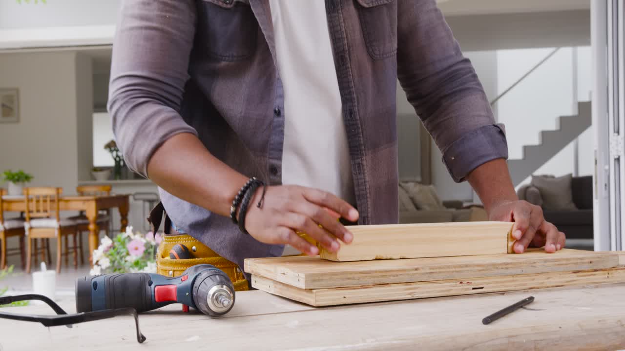 Uomo afroamericano che usa un trapano a batteria per avvitare viti in assi di legno su un patio, controllando l'allineamento.