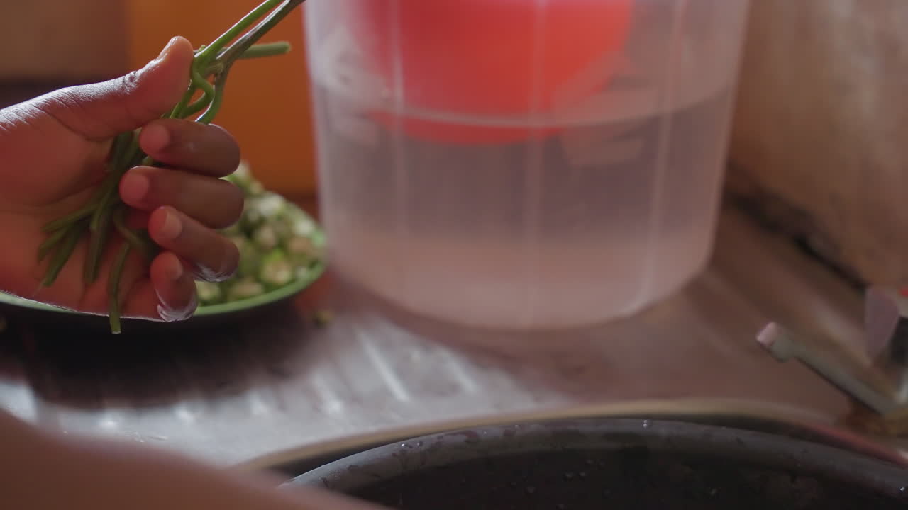 Mani in primo piano che lavano erbe aromatiche nel lavello, dita attente che staccano le foglie dai gambi sotto l'acqua tiepida del rubinetto, goccioline d'acqua che si disperdono, ciotola di verdure tritate sfocata sullo sfondo, preparazione in cucina domestica, pulizia tattile.