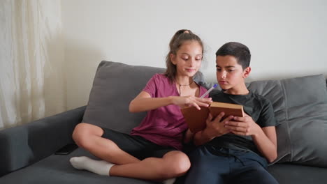 siblings sitting on couch studying together as girl flips to new book page with pen in hand while boy reacts with expressive face looking at page and mobile phone rests beside them
