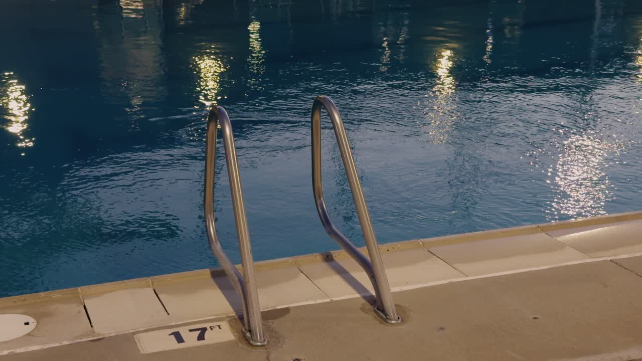 Calm blue pool water reflects lights at night beside metal pool ladder and deep-end marker