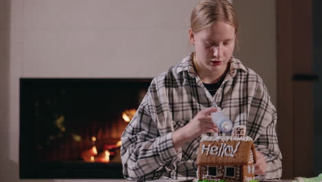 teenage girl decorating a gingerbread house by the fireplace