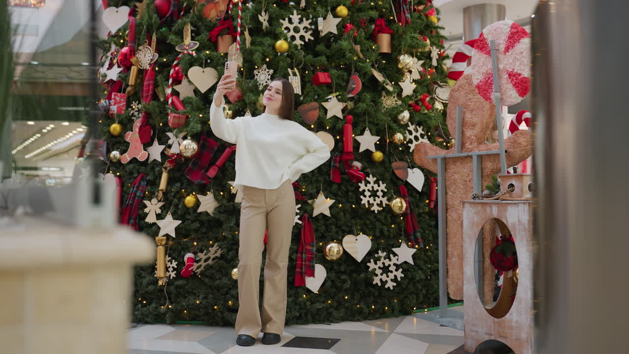 Young woman posing with smartphone to take selfie in front of beautifully decorated Christmas tree in mall, shoppers walking in background