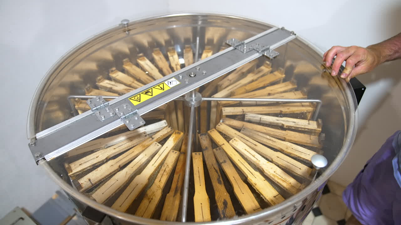 Metal centrifuge device for pumping out honey. Male hand closes apparatus with a transparent lid to avoid splatters.