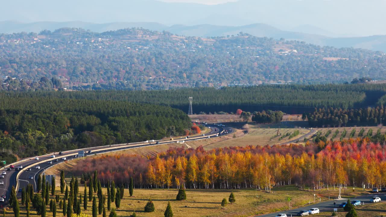 Late afternoon traffic flows steadily along Canberra’s Tuggeranong Parkway, surrounded by warm-toned autumn foliage and suburban rooftops in the distance.