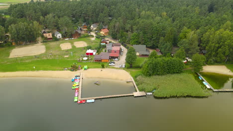 órbita-Aérea-Alrededor-De-Los-Complejos-Turísticos-Frente-Al-Lago-Hartowiec-Y-Edificios-De-Alquiler-Para-Vacaciones-De-Verano,-Campo-De-Voleibol-Y-Costa-En-Paisajes-Verdes,-Muelles-De-Madera-Con-Barcos-Atracados-En-El-Fondo-Del-Bosque