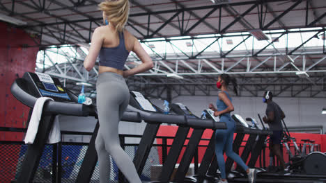 Two-fit-caucasian-women-and-fit-african-american-man-wearing-face-masks-running-on-treadmill-in-the-