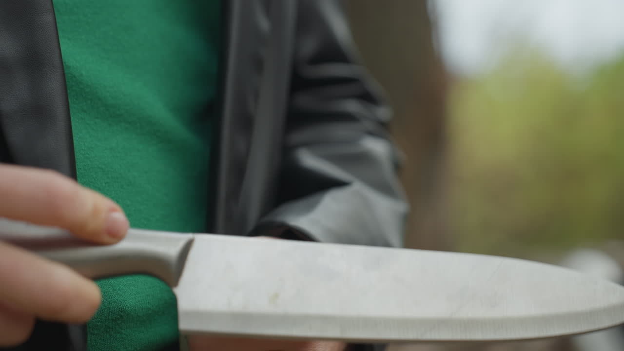 Mulher roqueira com jaqueta de couro segurando uma faca, close-ups de mãos ajustando a jaqueta e puxando a lâmina da bainha, suéter verde visível, floresta de outono ao fundo, clima cinematográfico tenso com textura