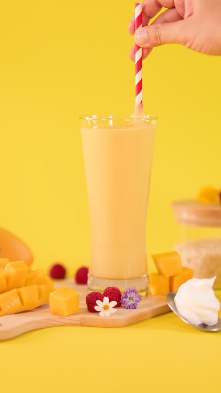 Primo piano di una mano che inserisce una cannuccia a strisce in un frullato freddo al mango su uno sfondo giallo brillante