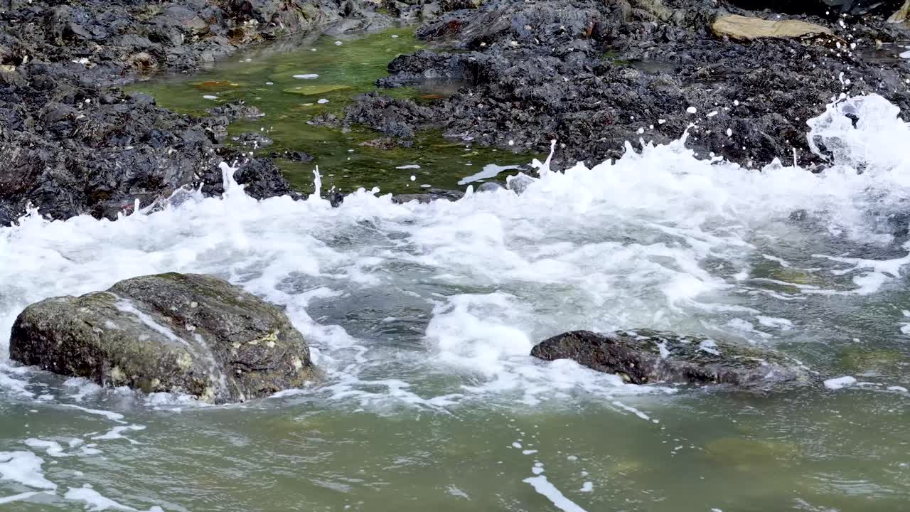 Coastal wave surges over rocks, creating whitewater and splashes in bright, natural daylight