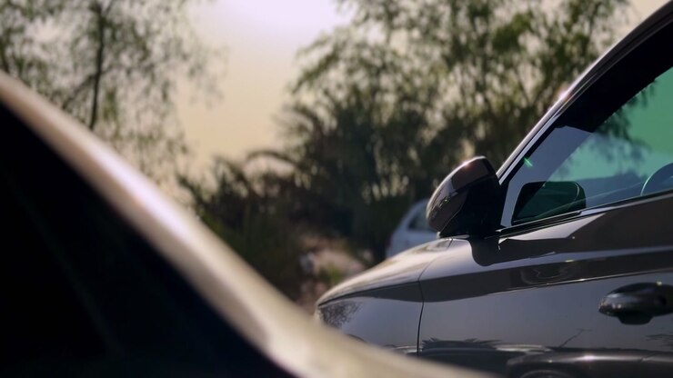 Primo piano di un'auto moderna grigio scuro parcheggiata all'aperto durante il giorno. La luce del sole si riflette sullo specchietto laterale e sui finestrini oscurati contro uno sfondo sfocato di alberi verdi e altri veicoli