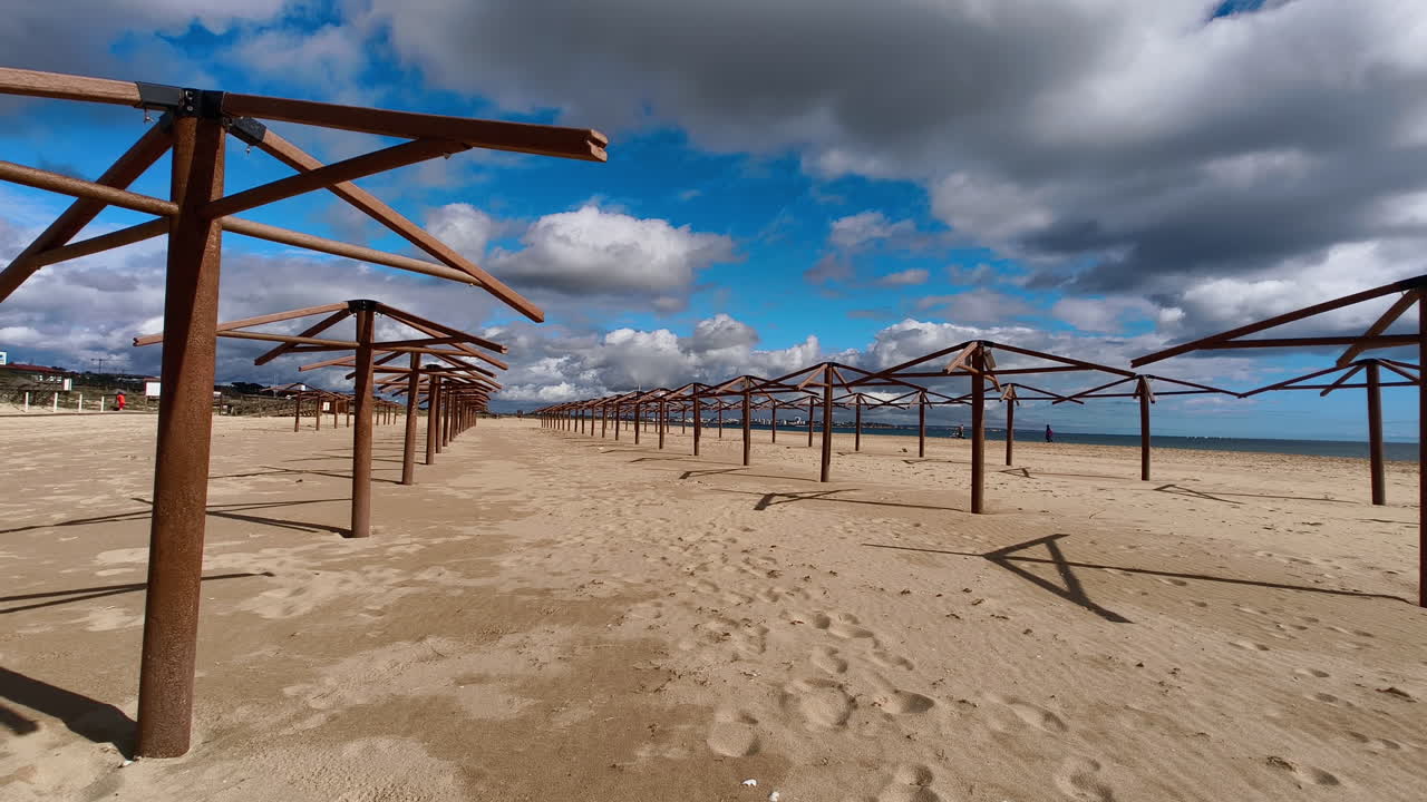 A long row of empty beach umbrellas stretches along a sandy beach. The sky is a bright blue with fluffy white clouds. In Lagos, meia praia, Portugal.