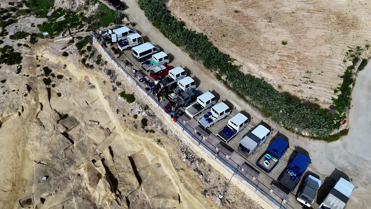 High Angle Drone Shot of Coastal Scuba Diving Expedition Fleet Parked on Rocky Limestone Cliffside During Bright Sunny Summer Day with Blue Vans and White Trucks Against Arid Mediterranean Landscape