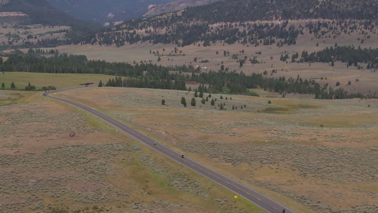 Two motorcyclists ride along a remote road through vast open fields in an aerial view