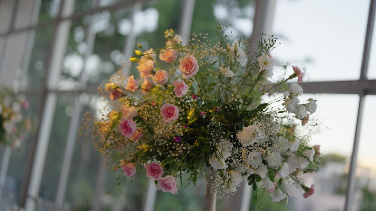 Soft slow‑motion footage focuses on a lush wedding centerpiece composed of pastel pink roses, white flowers and delicate greenery arranged in a tall vase, with the bright windows of a reception venue