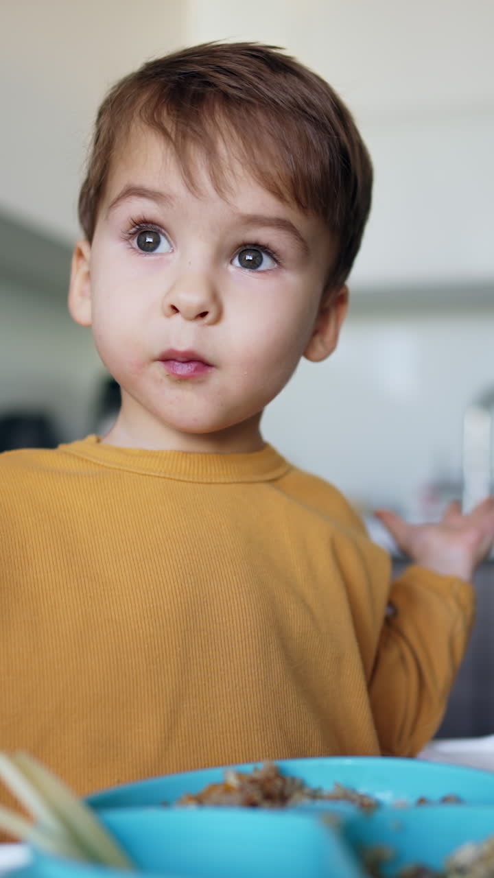 Lovely dark-haired baby boy sitting at high chair. Kid takes some food, places it on the plate and claps hands happily. Vertical video