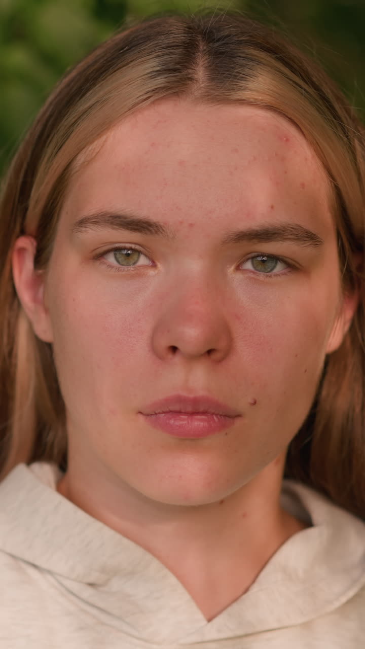 Young lady with bold expression, gazing directly at camera in natural setting, sunlight casts gentle shadows across her face, blending with lush green background
