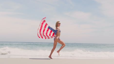 Seitenansicht-Einer-Kaukasischen-Frau-Mit-Wehender-Amerikanischer-Flagge,-Die-Am-Strand-Läuft-4k