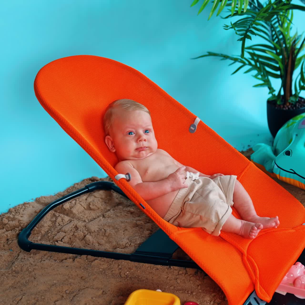 Lovely baby boy wearing shorts sits in the orange chair. Infant having rest on the improvised beach