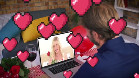 pixelated red heart icons floating over caucasian man waving while having a video call on laptop