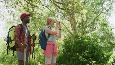 happy diverse couple hiking with backpacks and taking photos in park, slow motion