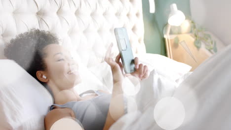 animation of white spots over biracial woman using smartphone in bed