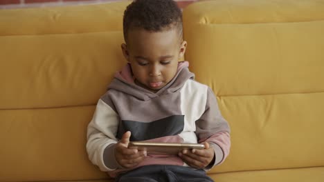 african little cute boy sitting on sofa using smartphone watching cartoons having fun