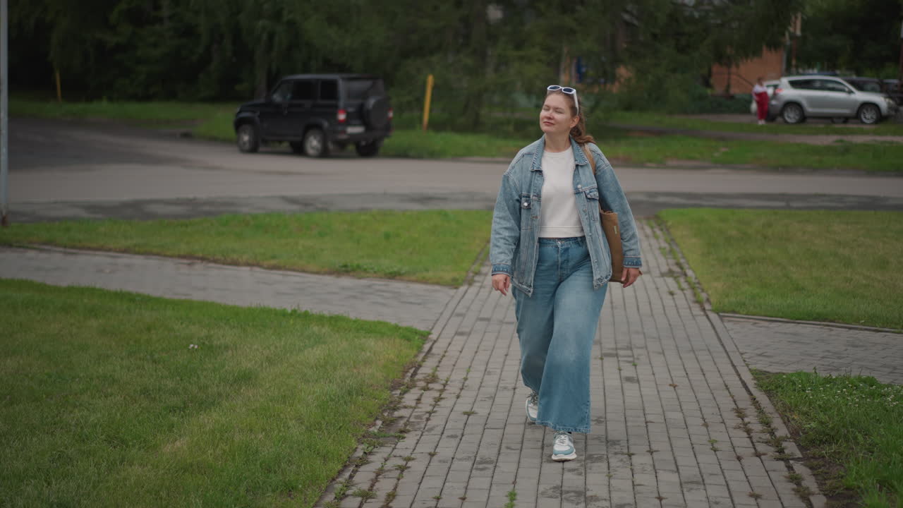 Caucasian woman walking on brick path, casual denim jacket and wide jeans, carrying satchel, confident stroll through suburban park with parked cars in background, soft overcast light, candid portrait