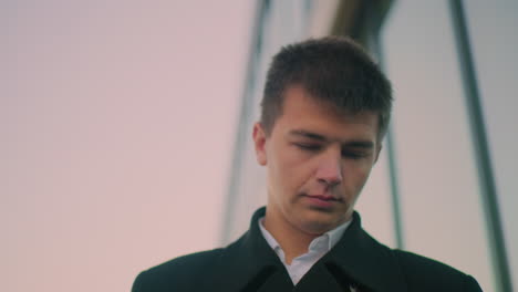 close-up of man in black coat using tablet focus near glass building, showing reflection of parked cars and blurred figures in glass at dusk