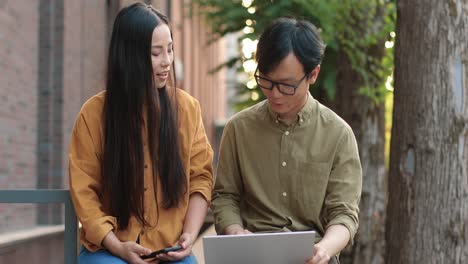 asian students sitting in the street and looking at the laptop while studying during a break and discussing something near the college