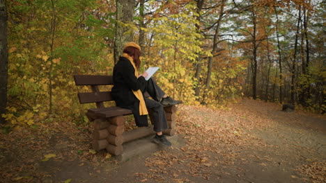 back view of lady sketching seated outdoors on wooden bench in solitude, surrounded by autumn foliage, immersed in creativity, wearing black coat and mustard yellow scarf