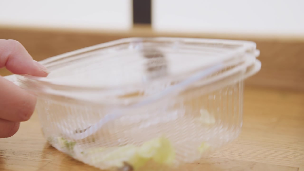 Plastic klapverpakking leeg achtergelaten op een houten aanrecht na de maaltijd, subtiele kruimels en een transparante bak tonen de nasleep van afhaalmaaltijden, minimaal keukenoppervlak en een rustig opruimmoment.