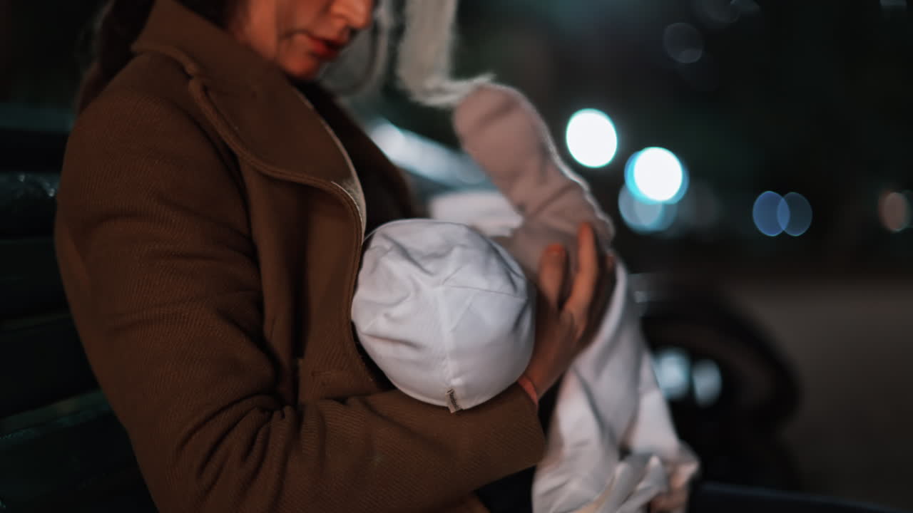 A thoughtful woman resting on a park bench under soft night lights, wrapped in a coat and holding a baby