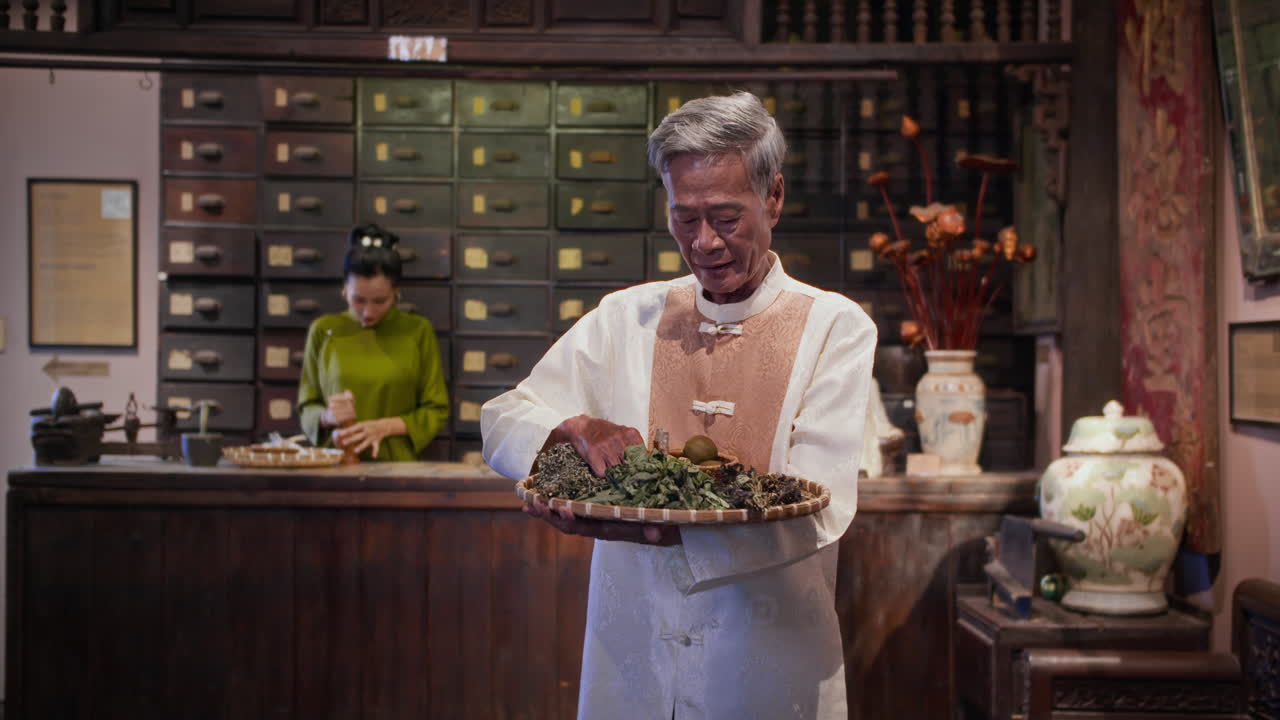 Asian Male Herbalist Holding Tray with Ingredients at Apothecary
