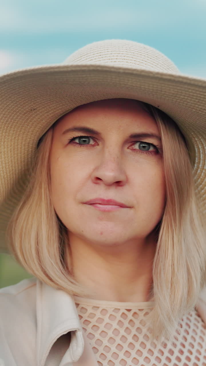 Elegant kvinde med solhat står udendørs, holder sin hat, mens hun eftertænksomt kigger ud i det fjerne; baggrunden viser et åbent landskab med træer, græs og en fredfyldt himmel.